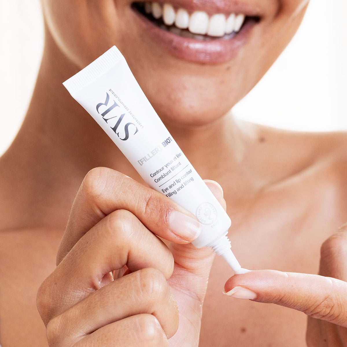 Una mujer sonriendo aplicando en una de sus manos el producto Filler Biotic de SVR en un fondo blanco