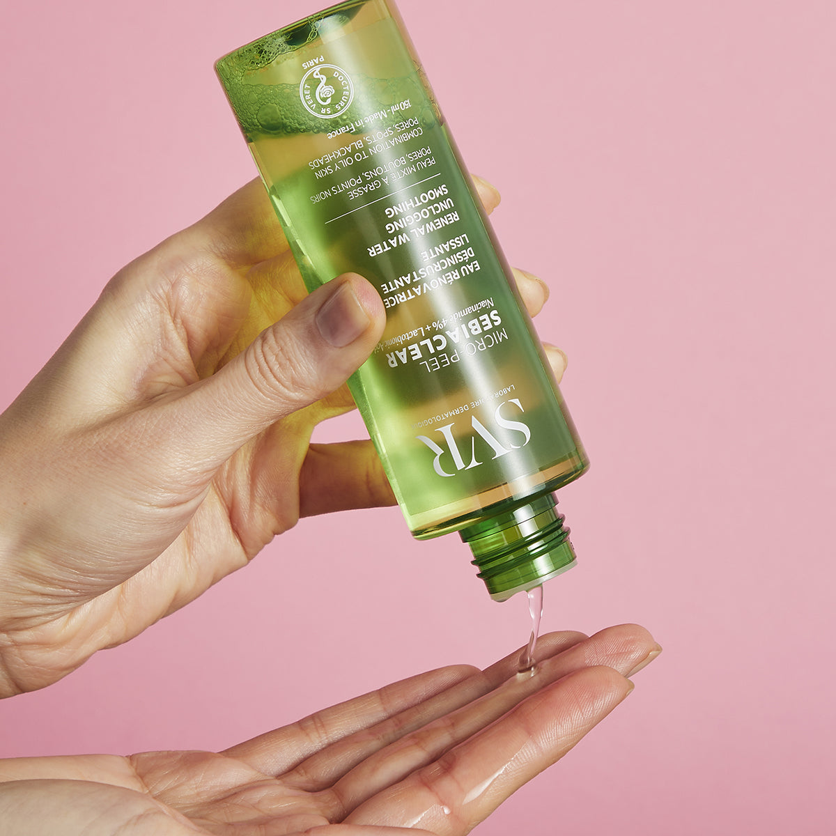 Frasco verde transparente de SVR Sebiaclear Micro-Peel vertiendo tónico líquido sobre una mano contra fondo rosa.