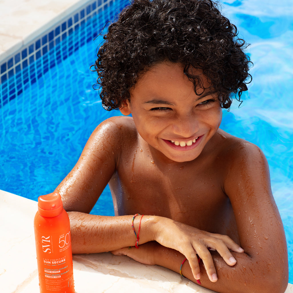 Niño moreno con rizos en la orilla de una piscina con Sun Secure Lait Crepitant al frente. 