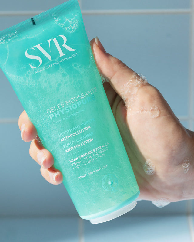 Una mano de tez clara mostrando el producto del gel PHYSIOPURE Gelee Moussante con espuma en la mano en un fondo azul
