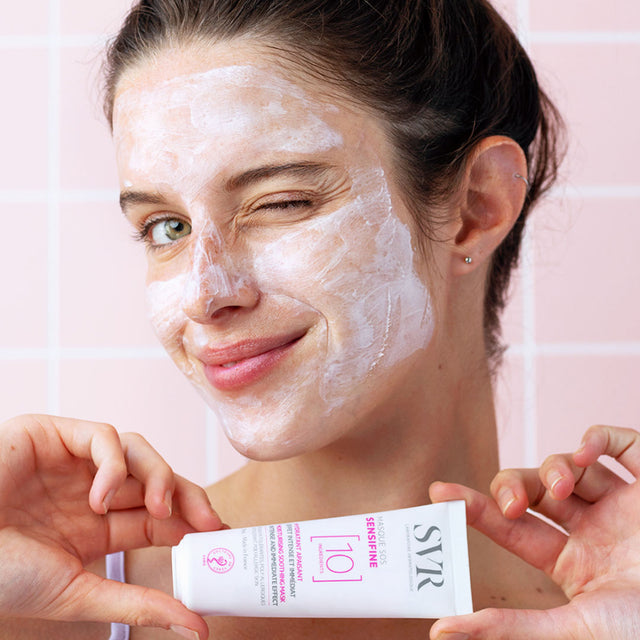Una mujer de tez clara con producto aplicado en su rostro de la crema hidratante Sensifine Masque SOS de SVR, en un fondo rosa claro 