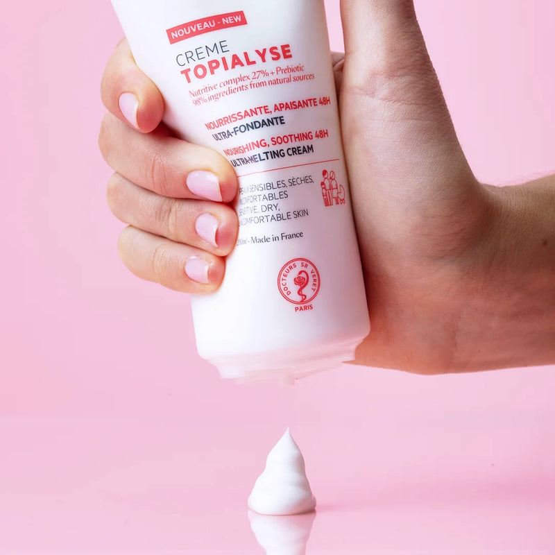 Hand squeezing a white tube of SVR Topialyse nourishing cream, dispensing a dollop of white lotion onto a pink surface.